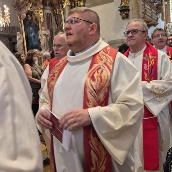 Priester stehen in einer Kirche, einer hält ein Buch, ein anderer mit einem Kreuz auf seiner Gewandung. Eine Laterne und religiöse Statuen schmücken den Hintergrund.
