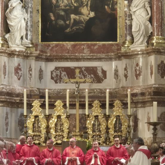 Geistliche stehen vor einem Altar mit Kerzen unter einem großen Gemälde in einer Marmorkirche.