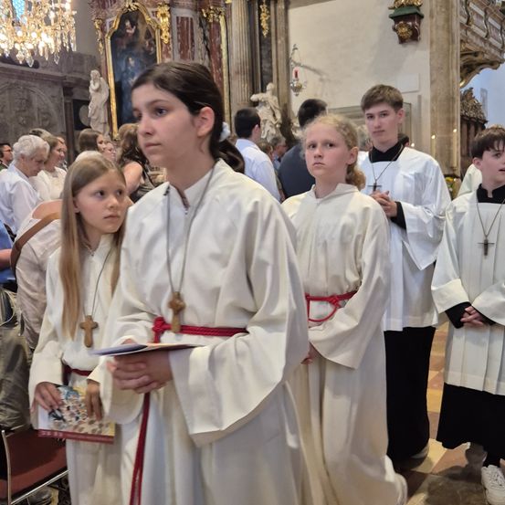 Eine Gruppe von Kindern in weißen Gewändern steht in einer Kirche, einige halten Bücher und andere haben die Hände gefaltet. Hinter ihnen beobachten Erwachsene von den Kirchenbänken aus. Im Hintergrund befinden sich eine Statue und ein Kronleuchter.