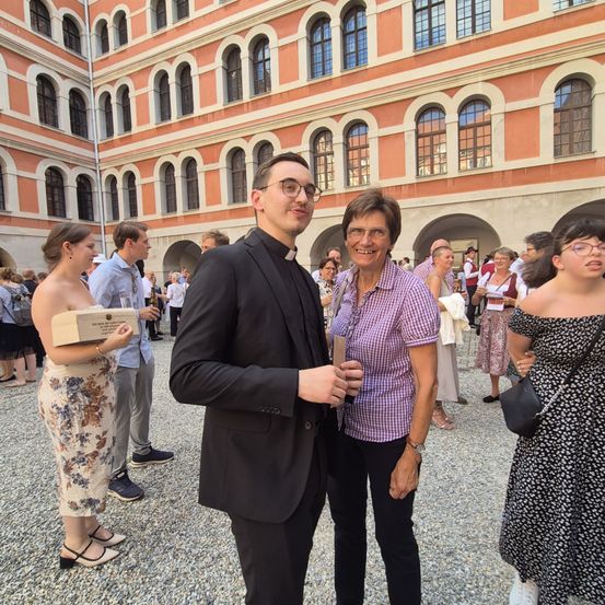 Eine Gruppe von Menschen steht vor einem prunkvollen Gebäude. Ein Mann in einem schwarzen Anzug hält ein Glas. Neben ihm steht eine lächelnde Frau in einem lila Oberteil. Andere Personen sind ebenfalls anwesend.