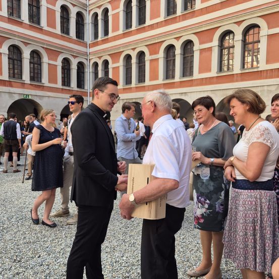 Eine Gruppe von Menschen steht in einem offenen Innenhof, möglicherweise bei einer gesellschaftlichen Veranstaltung. Ein Mann in einem schwarzen Anzug schüttelt die Hand eines älteren Mannes in einem weißen Hemd. Dahinter unterhalten sich mehrere andere Personen. Der Innenhof ist von einem alten Gebäude mit Bogenfenstern umgeben.
