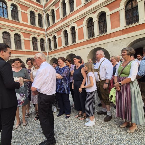 Eine Gruppe von Menschen steht vor einem historischen Gebäude, einige in traditioneller Kleidung. Sie scheinen einem Mann im Anzug zuzuhören, der zu ihnen spricht.