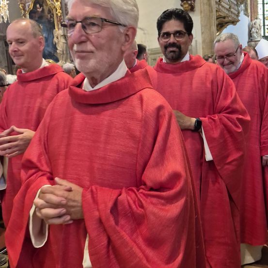 Mehrere Männer in roten Gewändern stehen in einer Kirche. Ein Mann mit Brille faltet seine Hände vor sich. Hinter ihm tragen zwei andere weiße Hüte.