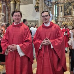 Zwei Männer in roten Roben stehen nebeneinander in einer Kirche und lächeln in die Kamera. Hinter ihnen sind weitere Personen anwesend, einige tragen Brillen und stehen in der Nähe des Altars.