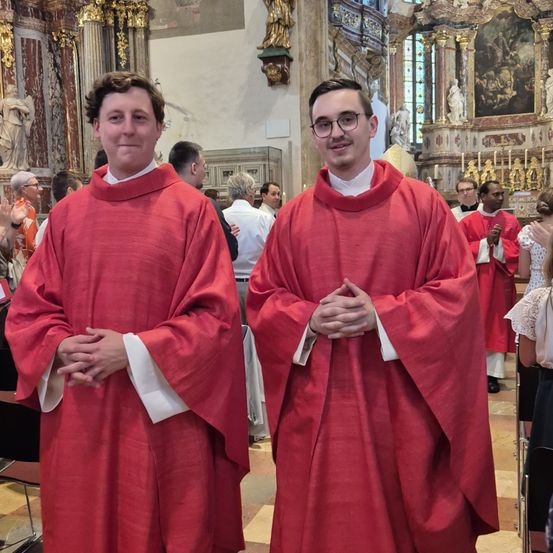 Zwei Männer in roten Roben stehen nebeneinander in einer Kirche und lächeln in die Kamera. Hinter ihnen sind weitere Personen anwesend, einige tragen Brillen und stehen in der Nähe des Altars.
