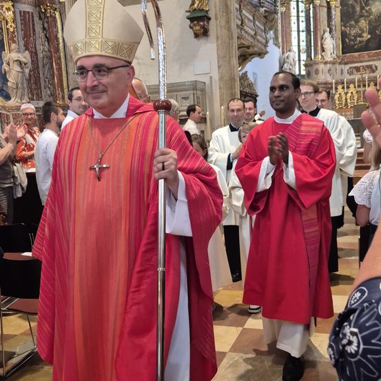 Ein Bischof steht in einer Kirche, hält einen Stab in der Hand und trägt einen roten Umhang und ein weißes Untergewand. Hinter ihm steht ein Mann in einem roten Umhang und einem weißen Untergewand und klatscht. Sie sind von Menschen umgeben, einige klatschen. Die Kirche hat einen gefliesten Boden, und eine Statue ist an der Wand montiert.