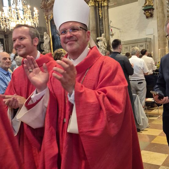 Mehrere Männer in roten Roben und Hüten stehen in einer Kirche. Ein Mann in einer roten Robe und einem weißen Hut gestikuliert mit den Händen.