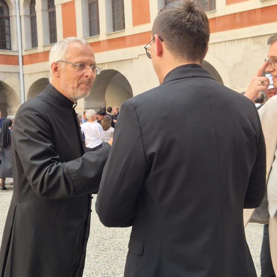 Ein älterer Mann in einem schwarzen Gewand und einer Brille spricht mit einem jüngeren Mann in einem schwarzen Anzug. Hinter ihnen gehen Menschen spazieren und unterhalten sich in der Nähe eines Gebäudes mit Rundbogenfenstern.