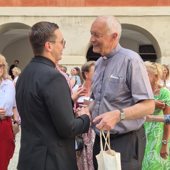Zwei Männer lächeln und schütteln sich die Hände in einer Außenumgebung. Der Mann in Grau hält eine weiße Tasche. Dahinter stehen mehrere Menschen, einige führen Gespräche. Eine Frau trägt eine Halskette.