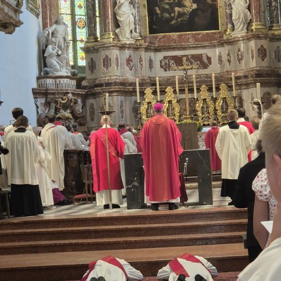 Priester in roten Gewändern versammeln sich in einer Kirche. Ein Priester in einem roten Gewand und einer rosa Mütze steht vor einer Kanzel. Kerzen und ein Gemälde schmücken den Altar.