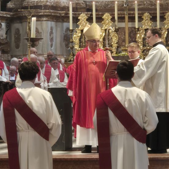 Eine religiöse Zeremonie findet in einer Kirche statt. Ein Bischof in einem roten Gewand hält ein Buch, während ein Assistent daraus liest.