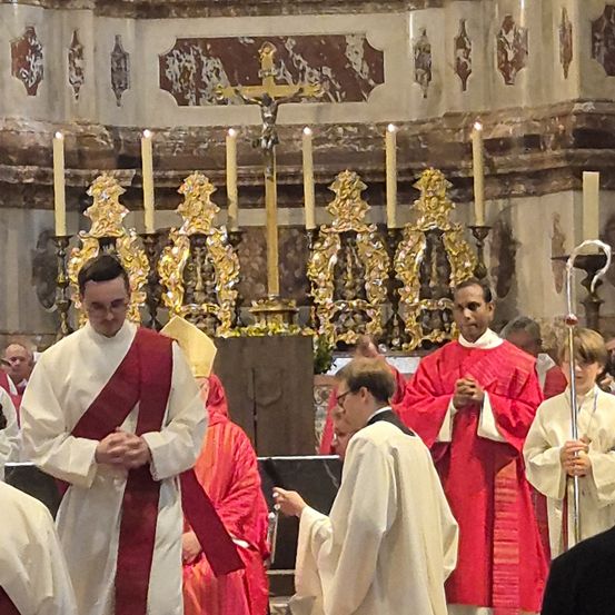 Geistliche in roten Gewändern und weißen Kaseln beten vor dem Altar. Kerzen sind angezündet und ein Kreuz steht in der Mitte.