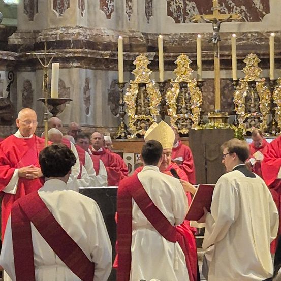Mehrere Priester in roten und weißen Gewändern sind in einer Kirche versammelt. Ein Bischof mit Mitra und Buch steht vor ihnen. Kerzen und ein Kreuz stehen auf dem Altar.