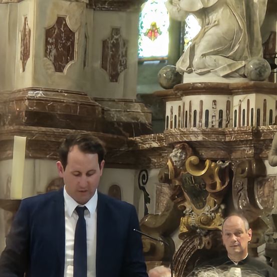 Ein Mann in Anzug steht vor einer Statue, möglicherweise in einer Kirche. Hinter ihm ist ein Priester und ein Altar mit einer Kerze.