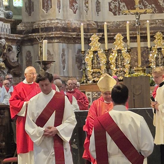 Eine Gruppe von Priestern in weißen und roten Gewändern, darunter einer mit einer goldenen Mitra, versammelt sich um einen Kirchenaltar. Kerzen und ein Kreuz stehen auf dem Altar.