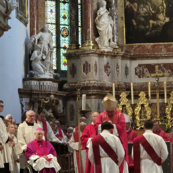 Eine religiöse Zeremonie mit einem Bischof in roten Gewändern, der eine Person im Rollstuhl segnet. Statuen und ein Buntglasfenster im Hintergrund.