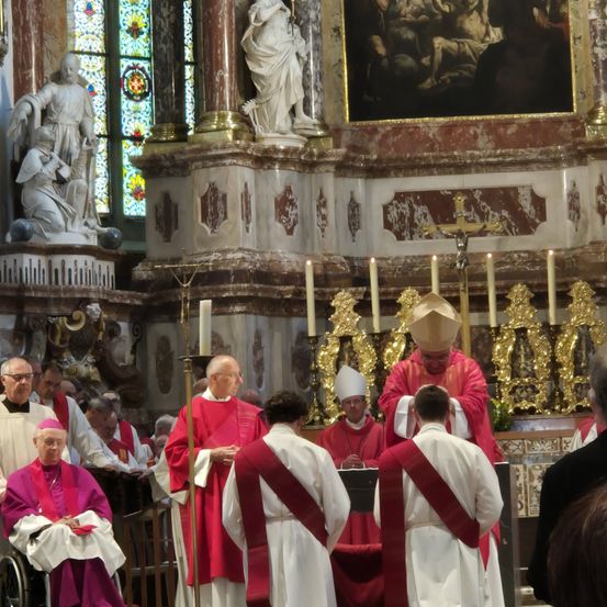 In einer Kirche versammeln sich mehrere Priester in roten und weißen Gewändern um einen Bischof in Rot. Kerzen und ein goldenes Kreuz schmücken den Altar. Statuen und Buntglasfenster sind vorhanden.
