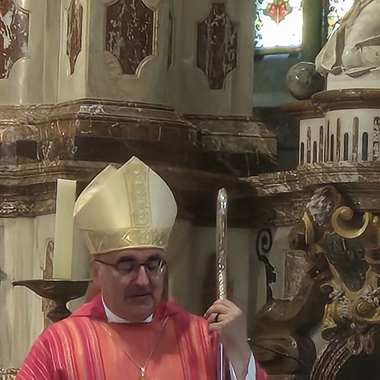 Ein Mann, der als Bischof gekleidet ist und eine Brille trägt, steht in einer Kirche mit einem goldenen Stab in der Hand. Die Kirche hat aufwendige Marmordesigns.