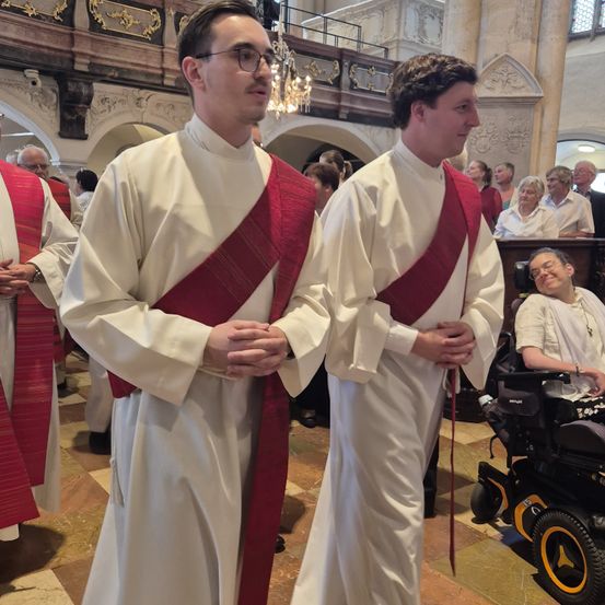 Zwei Priester in weißen Gewändern und roten Stolen stehen in einer Kirche. Hinter ihnen sitzen mehrere Menschen, darunter eine Person im Rollstuhl.