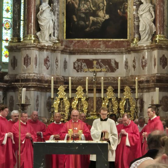 Priester in roten Roben stehen an einem Altar, umgeben von Kerzen und einem Gemälde darüber. Sie scheinen an einer religiösen Zeremonie teilzunehmen.