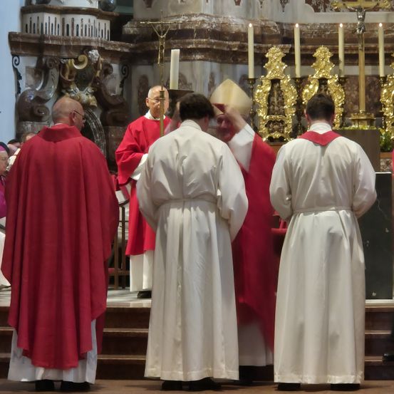 Mehrere Priester in roten und weißen Gewändern stehen in einer Kirche und nehmen an einer Zeremonie teil, bei der Kerzen auf dem Altar stehen.
