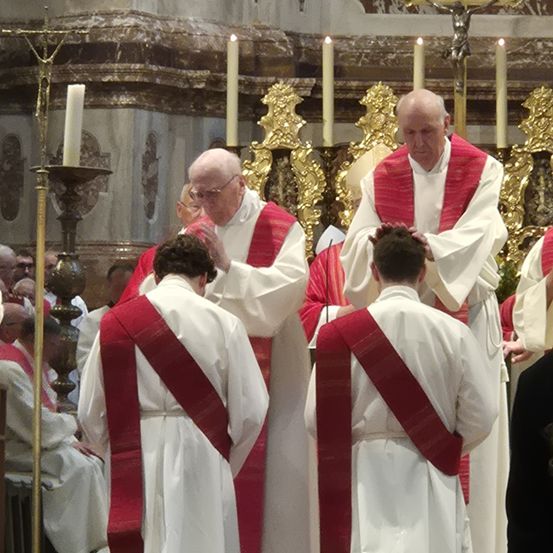 Priester in weißen Gewändern stehen in einer Kirche. Einige neigen ihre Köpfe. Sie stehen vor Kerzen auf Ständern. Eine Statue von Jesus Christus ist auf dem Altar.
