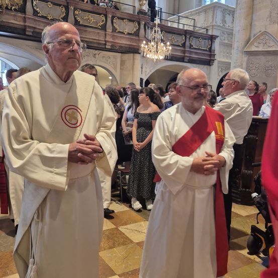 Zwei Priester stehen in einer Kirche während einer Zeremonie. Sie tragen weiße Gewänder, einer davon ein rotes Schultertuch. Eine Kronleuchter hängt über ihnen, und im Hintergrund sind Menschen versammelt.