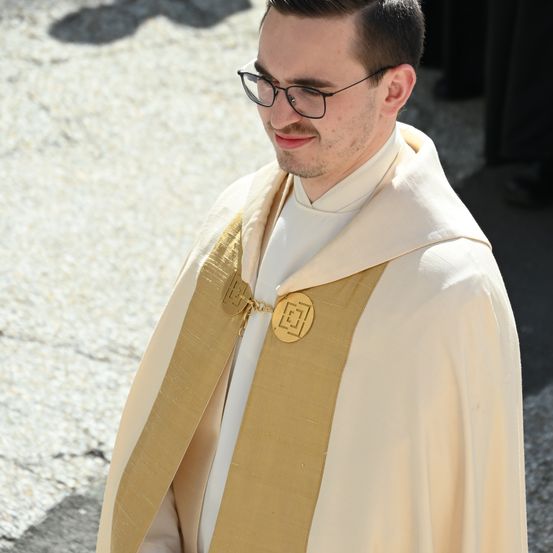 Ein Mann in einem weißen und goldenen Gewand, mit einer weißen Bluse und einer goldenen kreisförmigen Insignie auf seinem Gewand, steht auf einem Betonboden. Er trägt eine Brille.