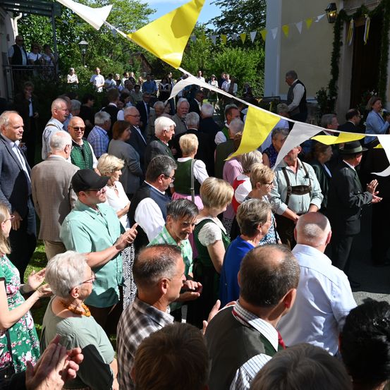 Eine Menschenmenge versammelt sich im Freien, einige klatschen, mit Wimpelketten und Fahnen. Im Hintergrund sind Bäume und ein Gebäude zu sehen.
