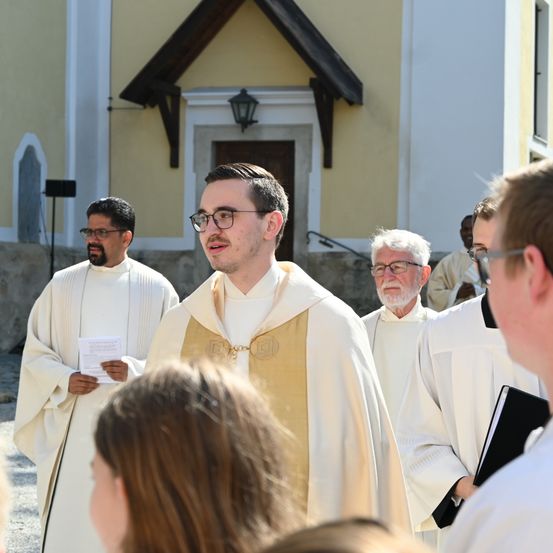 Eine Gruppe von Priestern in weißen Roben steht vor einer Kirche, einer hält ein Papier, ein anderer ein Buch, und die zentrale Figur trägt eine Brille und eine goldene Stola.