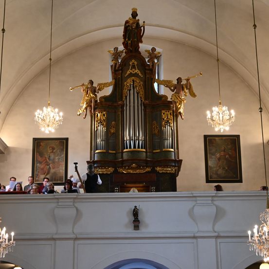 Ein Chor singt in einer Kirche mit einer großen, kunstvollen Orgel in der Mitte, flankiert von Gemälden und Kronleuchtern. Der Dirigent steht vor der Orgel mit einer Gruppe von Menschen darunter.