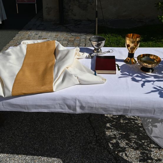 Ein weißer Stoff liegt auf einem Tisch mit einem Buch, Kelch und anderen religiösen Gegenständen. Schatten von nahegelegenen Objekten fallen auf den Tisch.