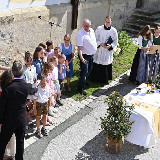 Ein Priester leitet eine Zeremonie im Freien, vor ihm steht eine Gruppe von Kindern und Erwachsenen vor einer Kirchenmauer. Ein Tisch mit einer Pflanze und religiösen Gegenständen steht in der Nähe.