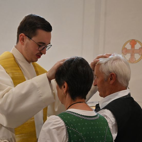 Ein Priester segnet eine Frau in einer Kirche, neben einem älteren Mann stehend, mit einem goldenen Kreuz im Hintergrund.