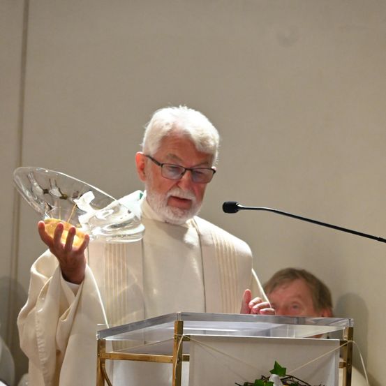Ein älterer Priester in einer weißen Robe hält eine klare Glasschale, möglicherweise während einer Zeremonie. Er steht vor einem Podium mit einem Mikrofon. Eine weitere Person steht hinter ihm und sieht ihm zu.