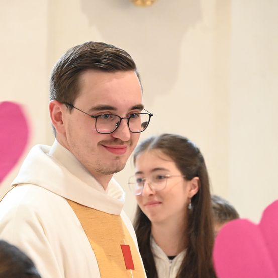 Ein lächelnder Mann in einer weißen und gelben Weste und einer Brille steht neben einer Frau mit langen Haaren und einer Brille. Sie sind von rosa Herzformen umgeben.
