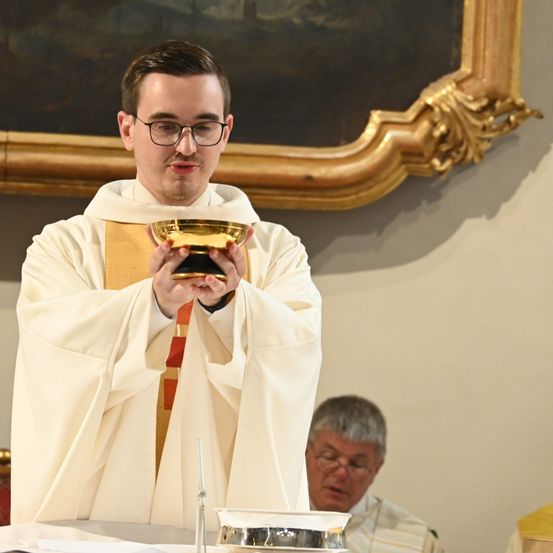 Ein Geistlicher in einer weißen Robe mit Brille hält einen goldenen Kelch, vermutlich während einer religiösen Zeremonie. Ein älterer Mann sitzt hinter ihm, ebenfalls mit Brille.