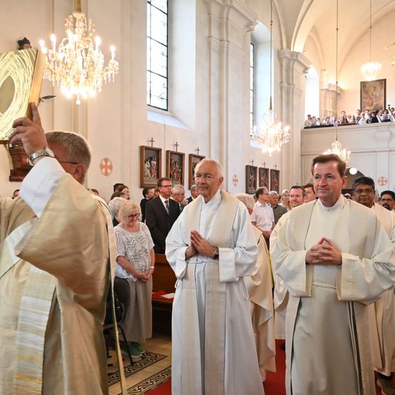 Priester in weißen Gewändern stehen in einer Kirche, einer hält ein Buch. Sie stehen vor dem Altar mit Kronleuchtern darüber. Zuschauer sitzen auf der Galerie.