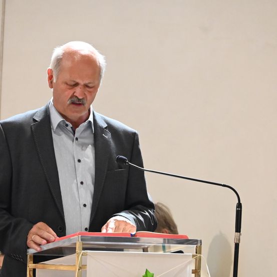 Ein älterer Mann mit einem Schnurrbart steht hinter einem Rednerpult mit Mikrofon. Er trägt ein graues Hemd und einen schwarzen Blazer. Ein grünes Blatt ist auf dem Podium.
