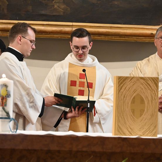 Drei Priester stehen an einem Altar, einer liest aus einem Buch, ein anderer hält eine Kerze und ein dritter steht neben einem hölzernen Kreuz.