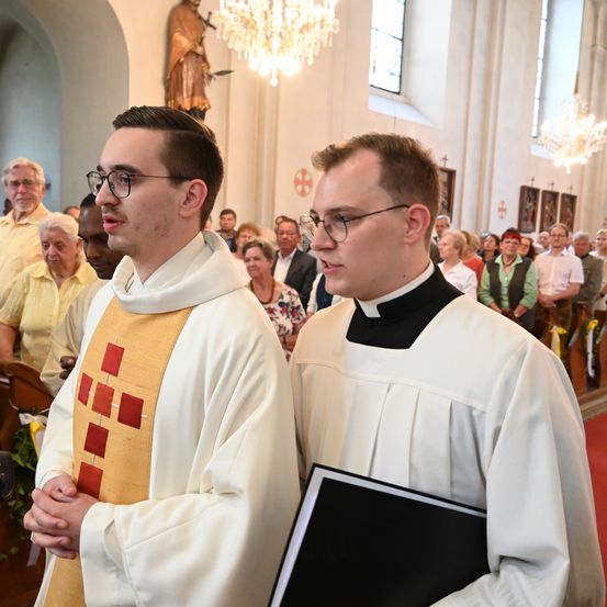 Zwei Priester stehen in einer Kirche mit kunstvollen Kronleuchtern und einer Statue im Hintergrund. Sie tragen beide eine Brille und weiße Gewänder.