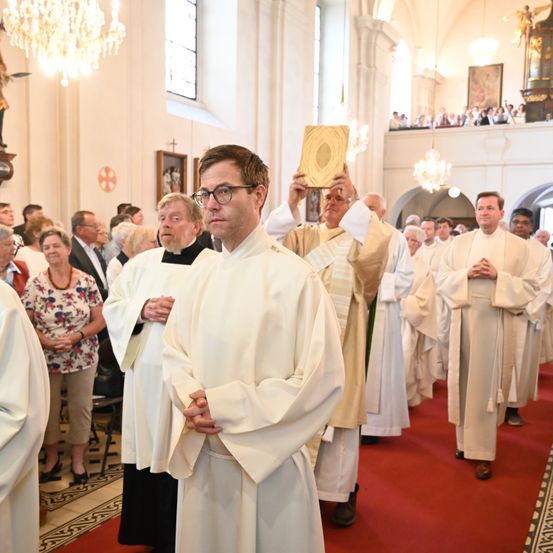 Eine Gruppe von Priestern in weißen Gewändern betet in einer Kirche. Der Priester in der Mitte hält ein Buch. Hinter ihnen sitzen Menschen auf den Bänken.