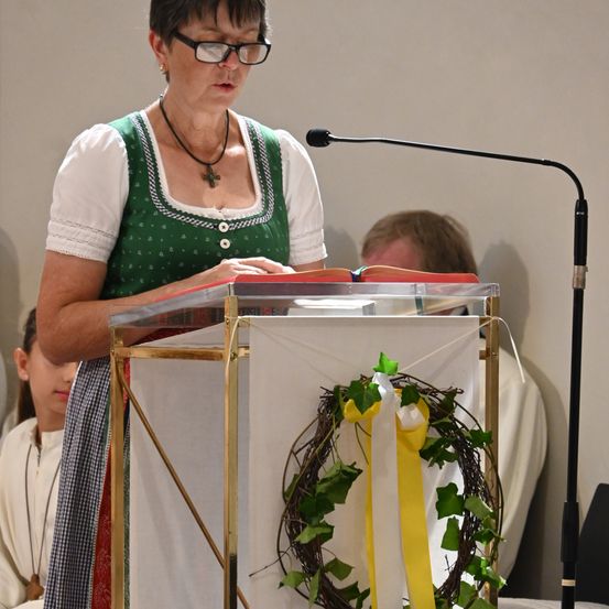 Eine Frau liest ein Buch an einem Pult, trägt eine Brille, eine Halskette und ein grünes und weißes Kleid. Hinter ihr ist eine Girlande mit grünen Blättern und einem gelben Band zu sehen.