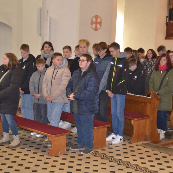 Eine Gruppe von Menschen steht in einer Kirche. Sie beten mit zusammengelegten Händen. Einige von ihnen tragen eine Brille. Hinter ihnen stehen Holzbänke und ein Kreuz an der Wand.