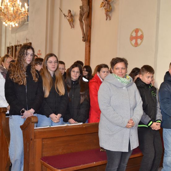 Mehrere Personen, möglicherweise Schüler, stehen in einer Kirche, einige tragen Jacken. Eine Frau steht getrennt und lächelt. Eine Holzbank mit einem roten Kissen steht hinter ihnen. Die Kirche hat dekorative Kreuze und religiöse Figuren.