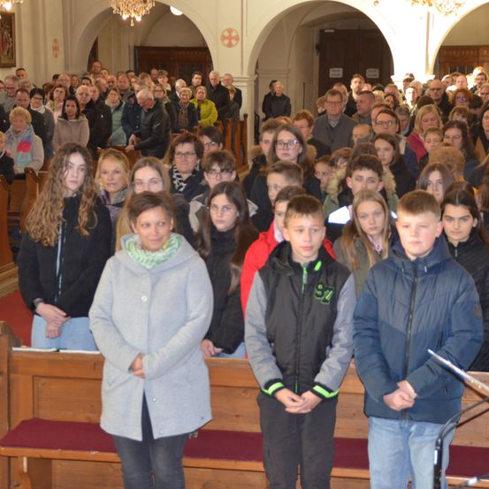 Eine Gruppe von Menschen steht in einer Kirche, möglicherweise während eines Gottesdienstes. Eine Frau in einem grauen Mantel steht vorne. Hinter ihr sitzen und stehen Menschen. Es gibt Kronleuchter an der Decke.
