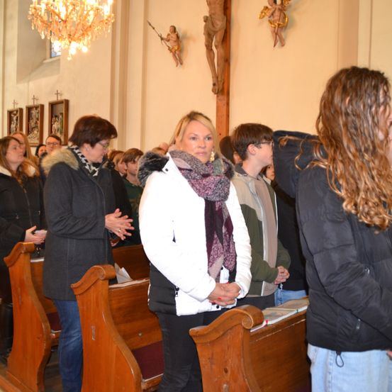 Eine Gruppe von Menschen steht in einer Kirche, gekleidet in Winterkleidung. Eine Frau in einer weißen Jacke steht in der Mitte, und eine Person mit langen Haaren betet. Die Kirche hat Holzbänke, eine Kristallleuchter und religiöse Ikonen an der Wand.