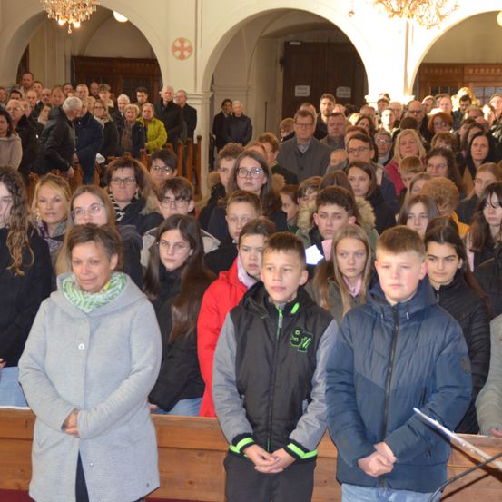 Eine große Menschenmenge versammelt sich in einer Kirche, alle in Winterkleidung. Einige stehen, andere sitzen auf Bänken. Die Kirche hat bogenförmige Fenster und Holzbänke.