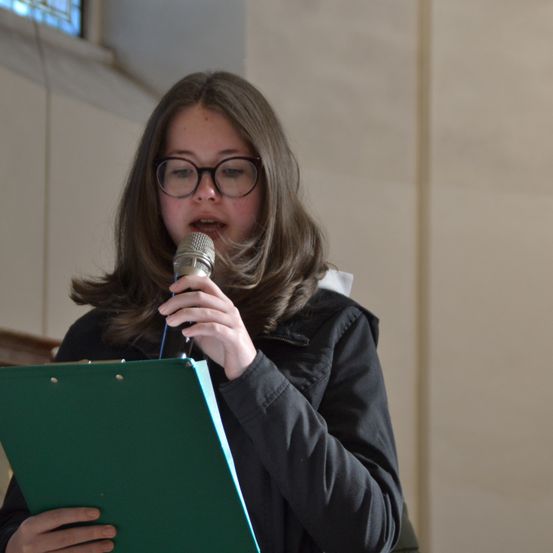 Eine Frau mit Brille hält ein Mikrofon in der rechten Hand und eine grüne Mappe in der linken. Sie blickt nach unten und scheint etwas zu lesen. Der Raum ist schwach beleuchtet.