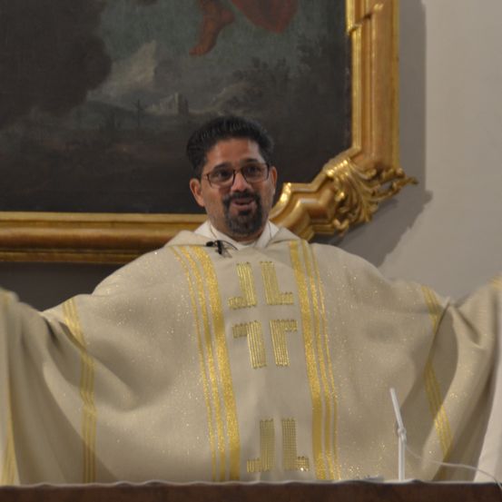 Ein Priester steht an einem Rednerpult und trägt einen weißen und goldenen Gewand mit goldener Stickerei. Hinter ihm schmückt ein gerahmtes Gemälde die Wand.